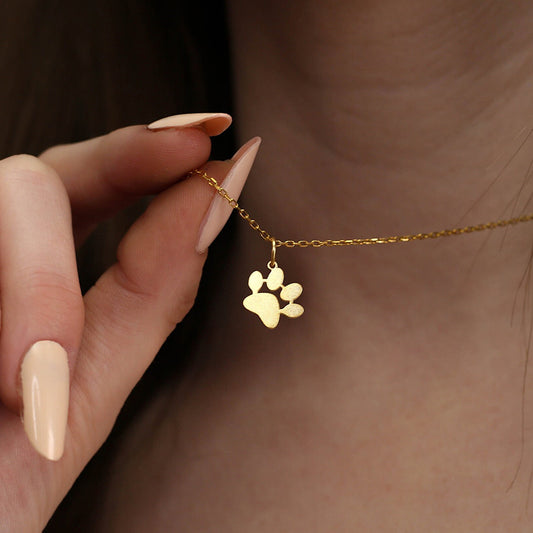 A close-up lifestyle shot of a person holding a delicate lilyah jewelry's 18k gold plated chain necklace with a small paw print pendant. The person has long, manicured fingernails painted in a light nude shade, and the background is a soft-focus view of their neck and hair.