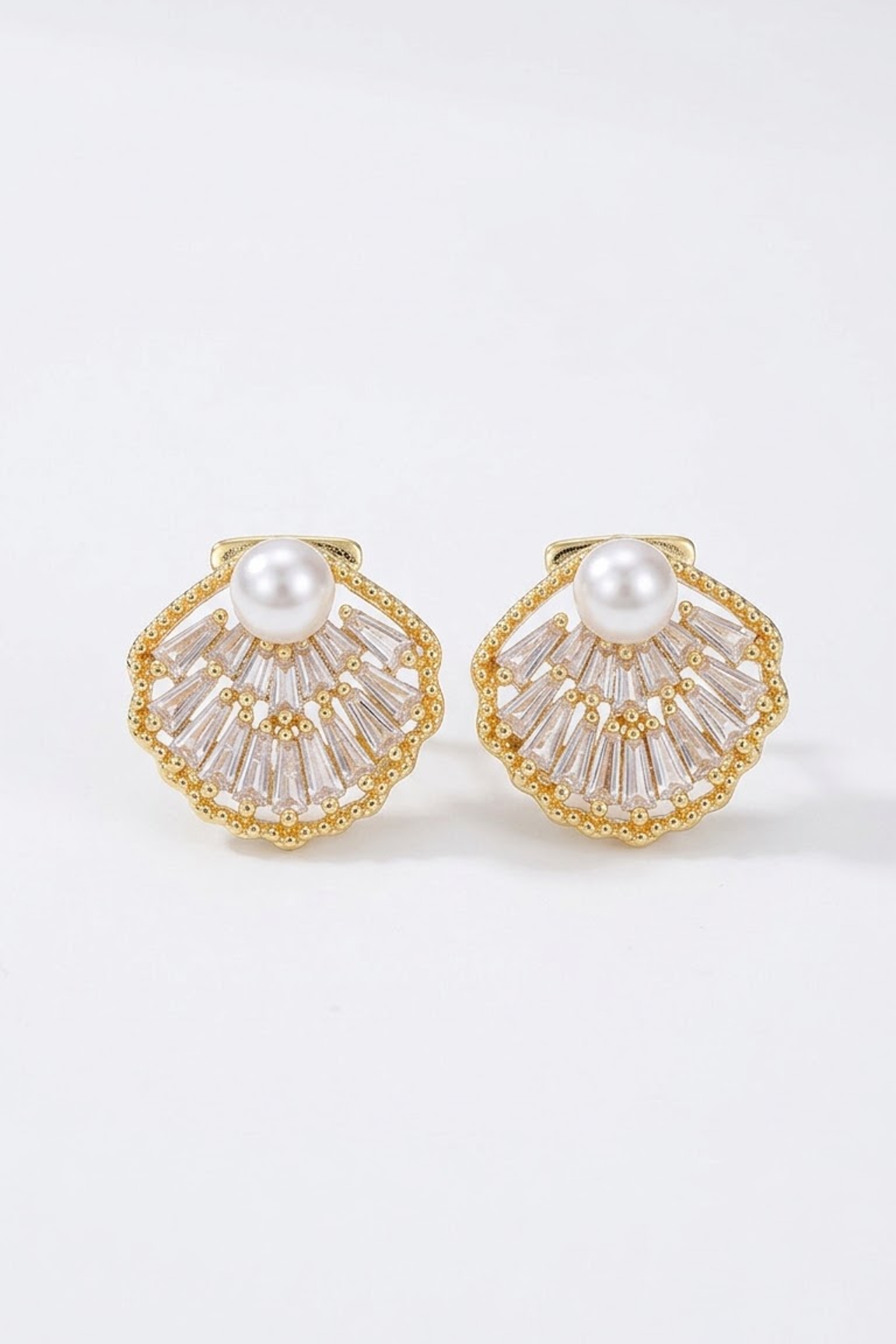 A pair of 18k gold-toned shell-shaped earrings with a white pearl at the center and clear baguette crystals radiating from the edges, shown on a clean white background.