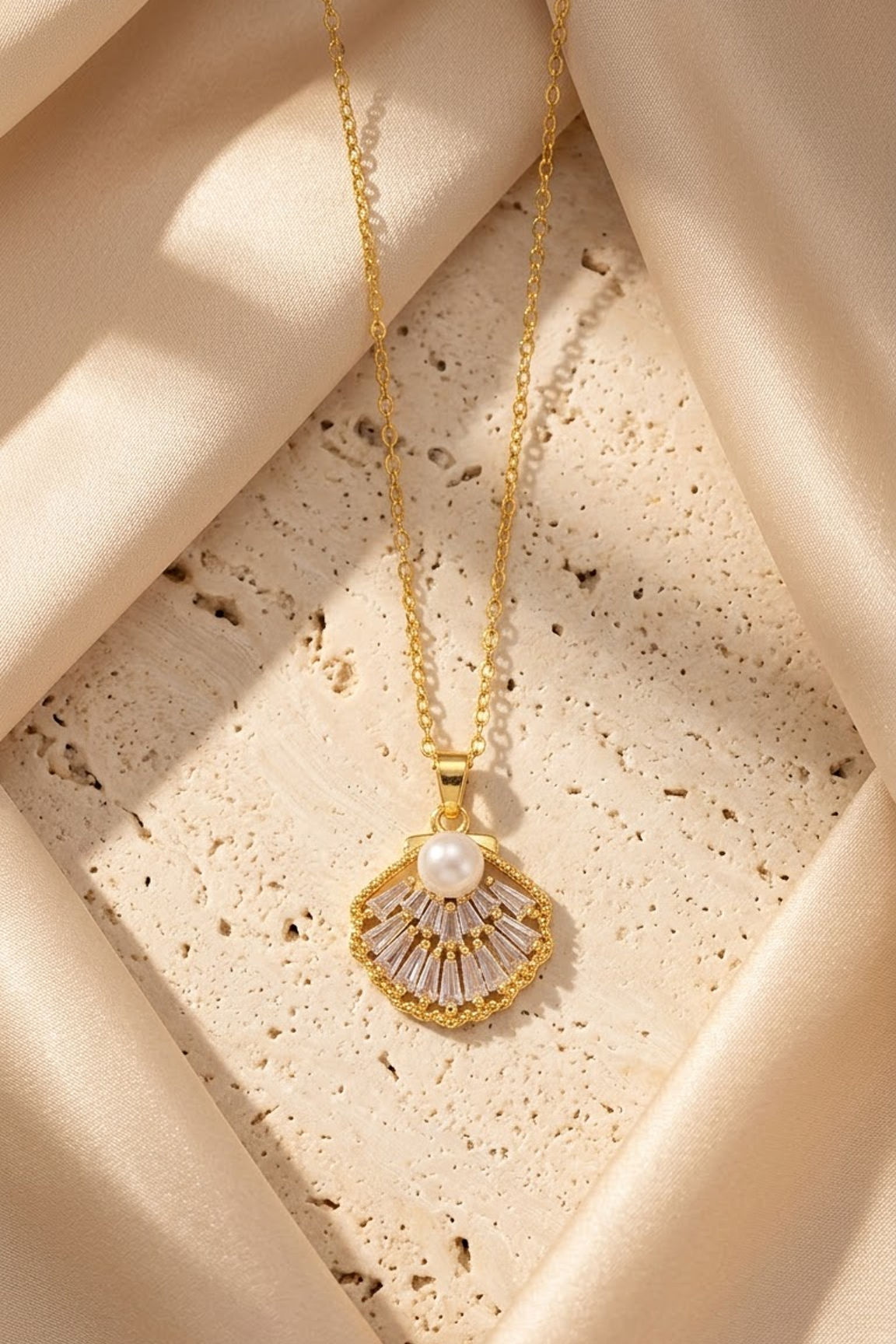 Lilyah Jewelry's 18k gold plated pendant necklace with a scalloped shell design, featuring a central white pearl and clear stones, draped elegantly over a textured beige stone surface with cream satin fabric.