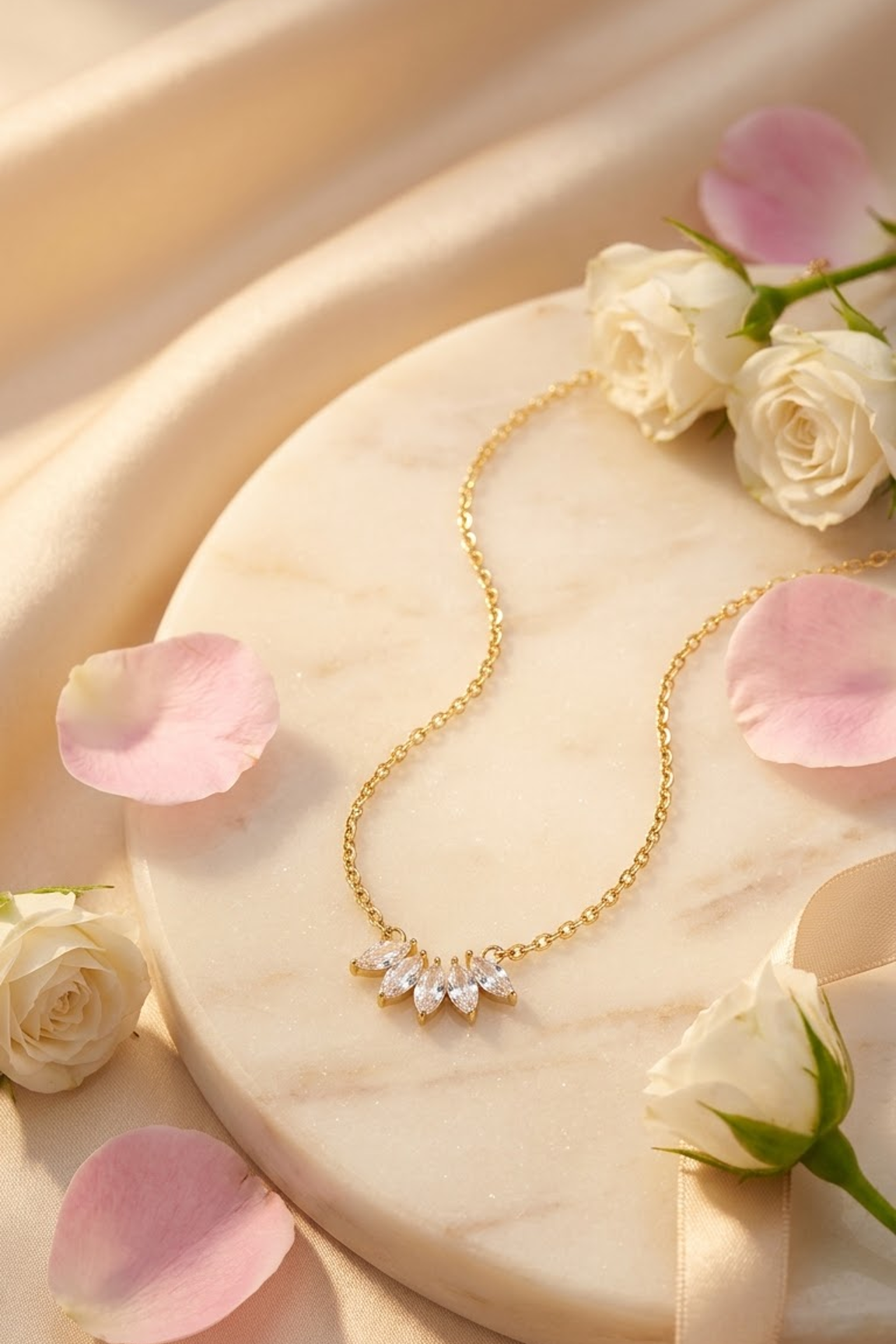 A delicate 18k gold plated chain necklace with a small floral cluster pendant set with clear rhinestones from the brand Lilyah Jewelry, displayed on a round marble tray with white roses and pink petals.