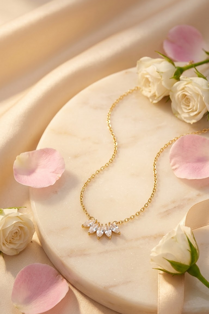 A delicate 18k gold plated chain necklace with a small floral cluster pendant set with clear rhinestones from the brand Lilyah Jewelry, displayed on a round marble tray with white roses and pink petals.