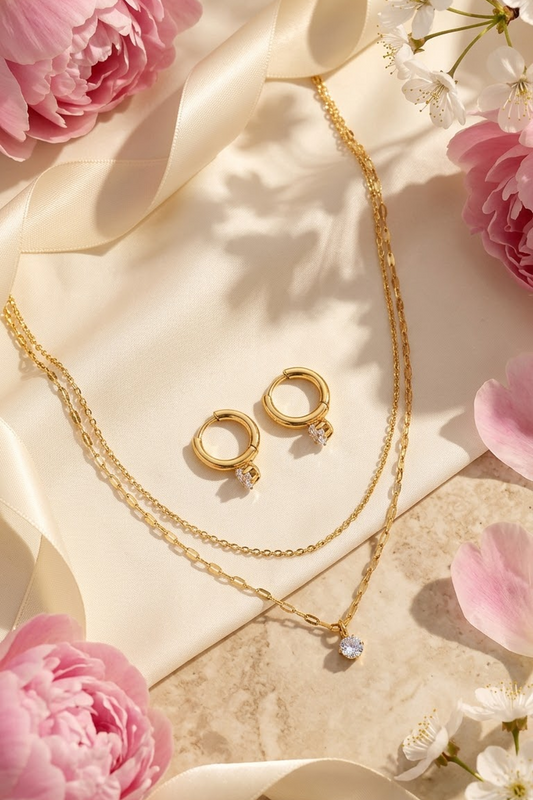 Lilyah Jewelry's 18k Gold plated jewelry set including a layered chain necklace with a small clear stone pendant, a pair of gold hoop earrings with matching clear stone accents, arranged on cream satin with pink flowers and ribbon around them.
