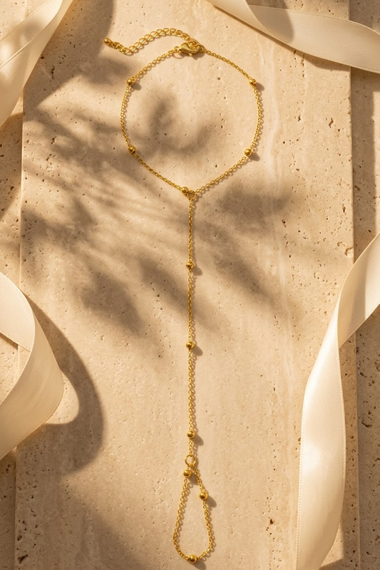 Lilyah Jewelry's delicate 18k gold plated bracelet cum ring with small bead details, laid flat on a textured beige stone surface with soft cream ribbons and leaf shadows.