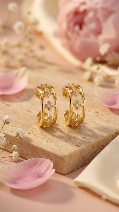 A pair of Lilyah Jewelry's 18k gold plated ear cuff earrings with floral and geometric cutout details, displayed on a beige stone slab with pink petals and soft flowers in the background.
