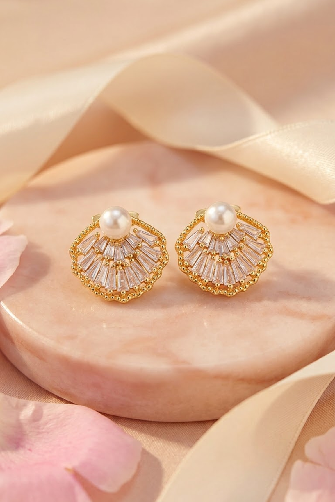 Lilyah Jewelry's 18k gold plated shell shaped stud earrings with clear crystal facets and a central white pearl, displayed on a round pink marble slab with soft satin ribbon and pink petals.