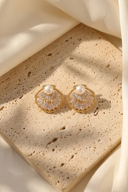 Lilyah Jewelry's pair of gold-toned shell-shaped earrings with clear zircons and a central white pearl, displayed on a beige stone slab with draped cream fabric.