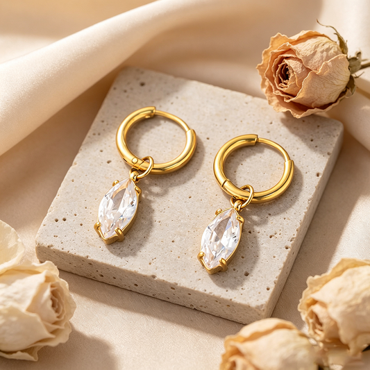A pair of Lilyah Jewelry's 18k gold plated hoop earrings with marquise-cut clear zircon drops, displayed on a textured stone slab with dried roses.