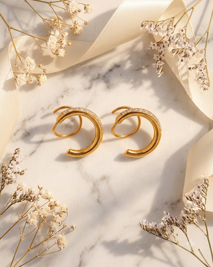 A pair of 18k gold plated spiral shaped hoop earrings from lilyah jewelry with small clear zircons along the front, displayed on a white marble surface with cream ribbon and dried flowers.