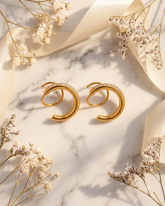 A pair of 18k gold plated spiral shaped hoop earrings from lilyah jewelry with small clear zircons along the front, displayed on a white marble surface with cream ribbon and dried flowers.